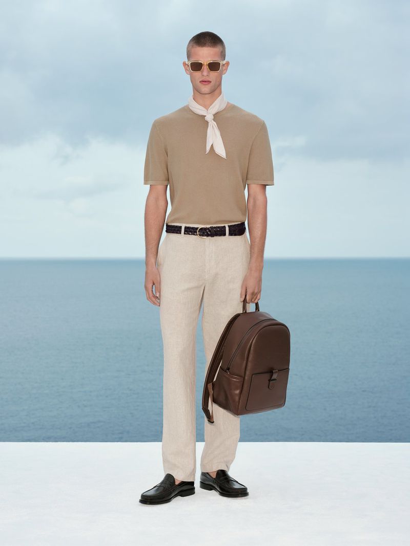 Man wearing beige knit top, light trousers, neck scarf, and holding a brown leather backpack against an ocean backdrop.