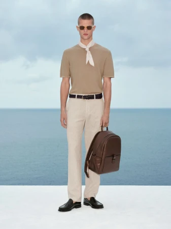 Man wearing beige knit top, light trousers, neck scarf, and holding a brown leather backpack against an ocean backdrop.