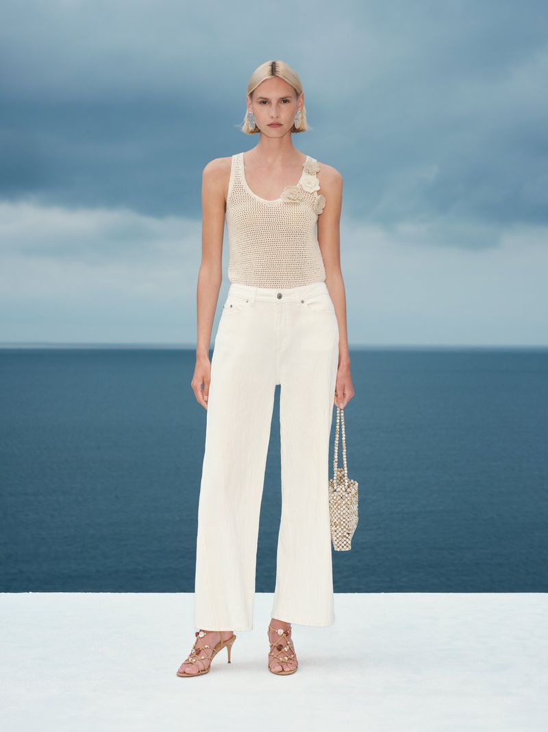 Blonde female model wearing cream knitted top and white trousers holding beaded handbag by the ocean
