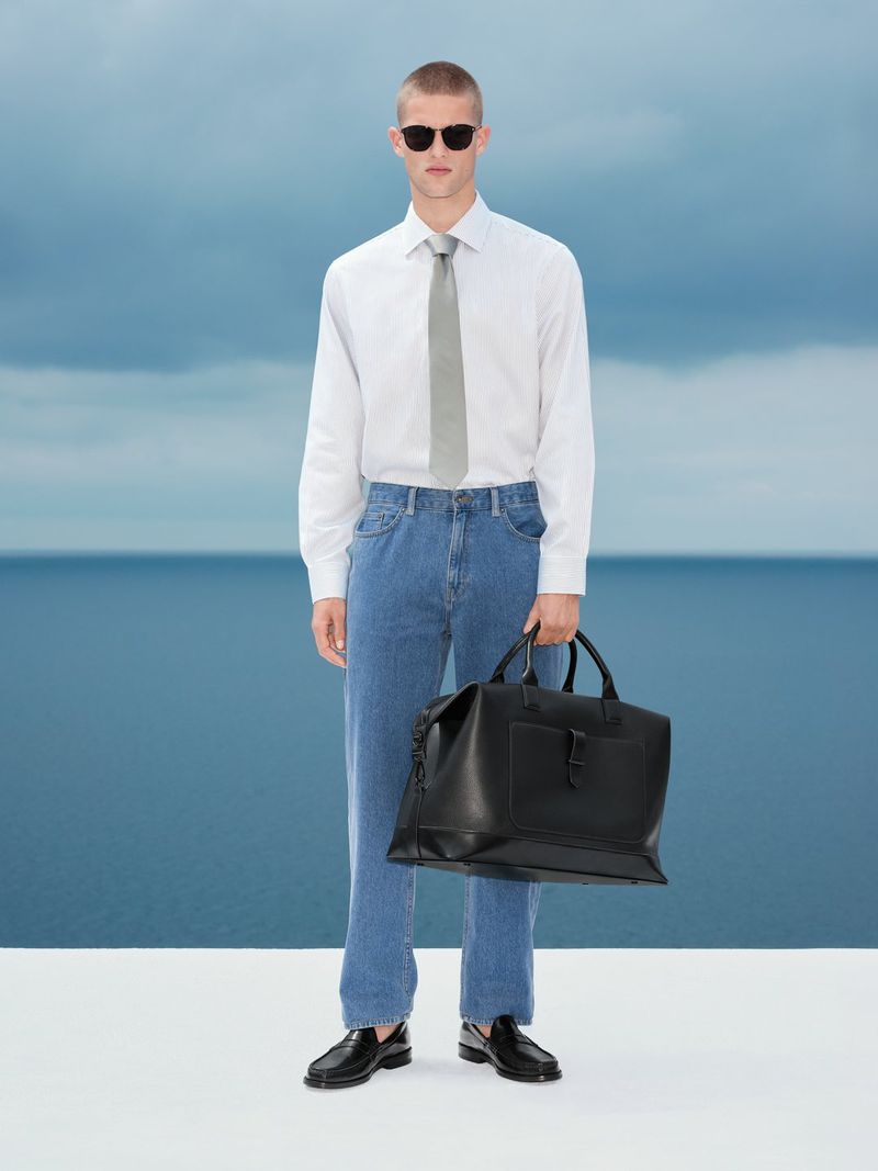 Model wearing a white shirt and blue jeans holding a black duffel bag against a coastal backdrop