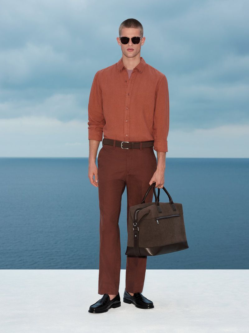 Model wearing a rust-colored shirt and brown trousers holding a dark travel bag against an ocean background.