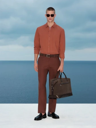 Model wearing a rust-colored shirt and brown trousers holding a dark travel bag against an ocean background.