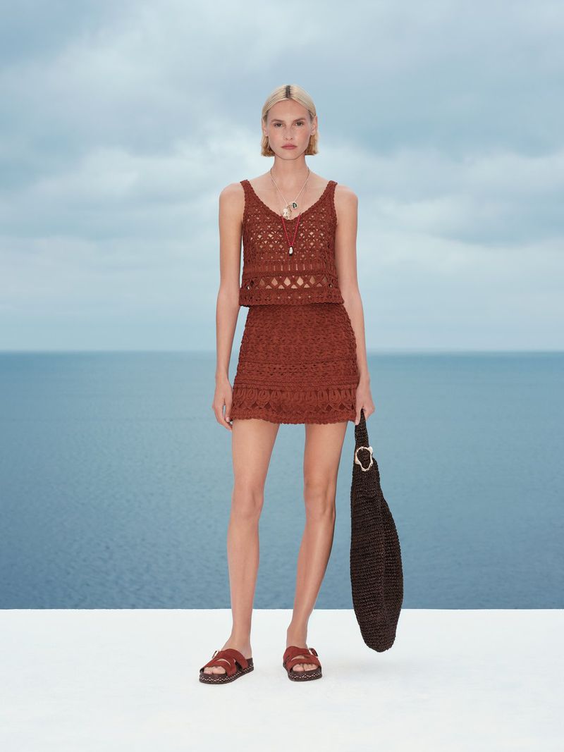 Model in a brown crochet knit top and skirt holding a dark woven bag against an ocean background