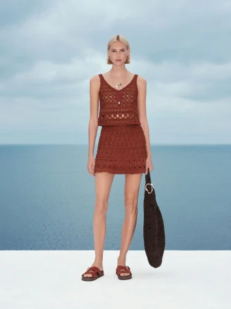 Model in a brown crochet knit top and skirt holding a dark woven bag against an ocean background