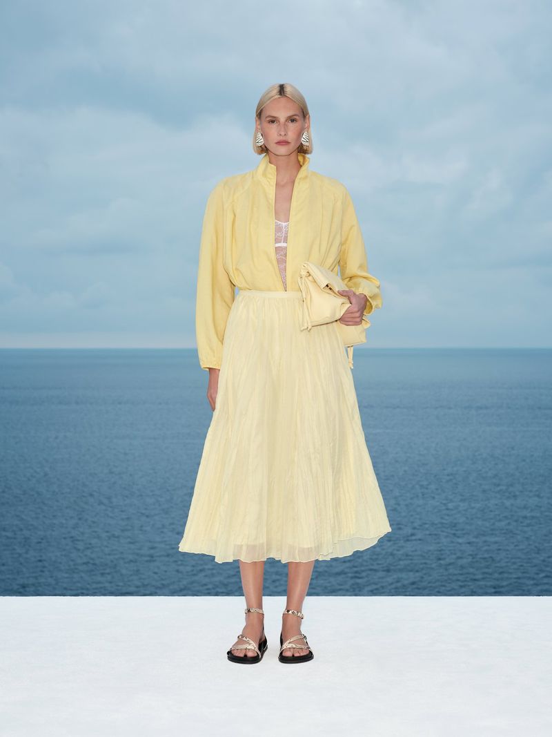 Model wearing yellow long-sleeve blouse and pleated skirt holding a clutch bag against a sea view.