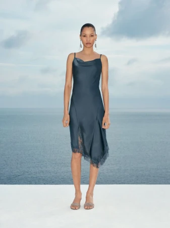 Model wearing a dark blue satin slip dress standing against an ocean and cloudy sky backdrop