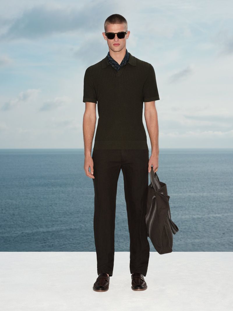 Man wearing sunglasses, dark polo shirt, and trousers holding a bag against an ocean and sky background for Marks & Spencer