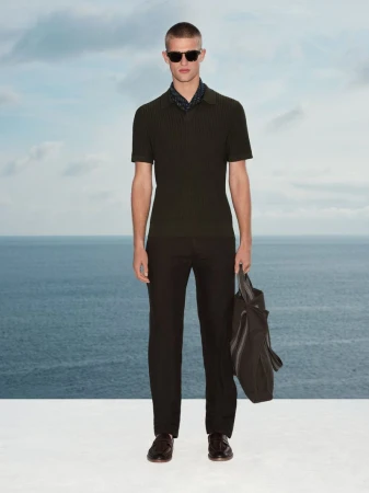 Man wearing sunglasses, dark polo shirt, and trousers holding a bag against an ocean and sky background for Marks & Spencer