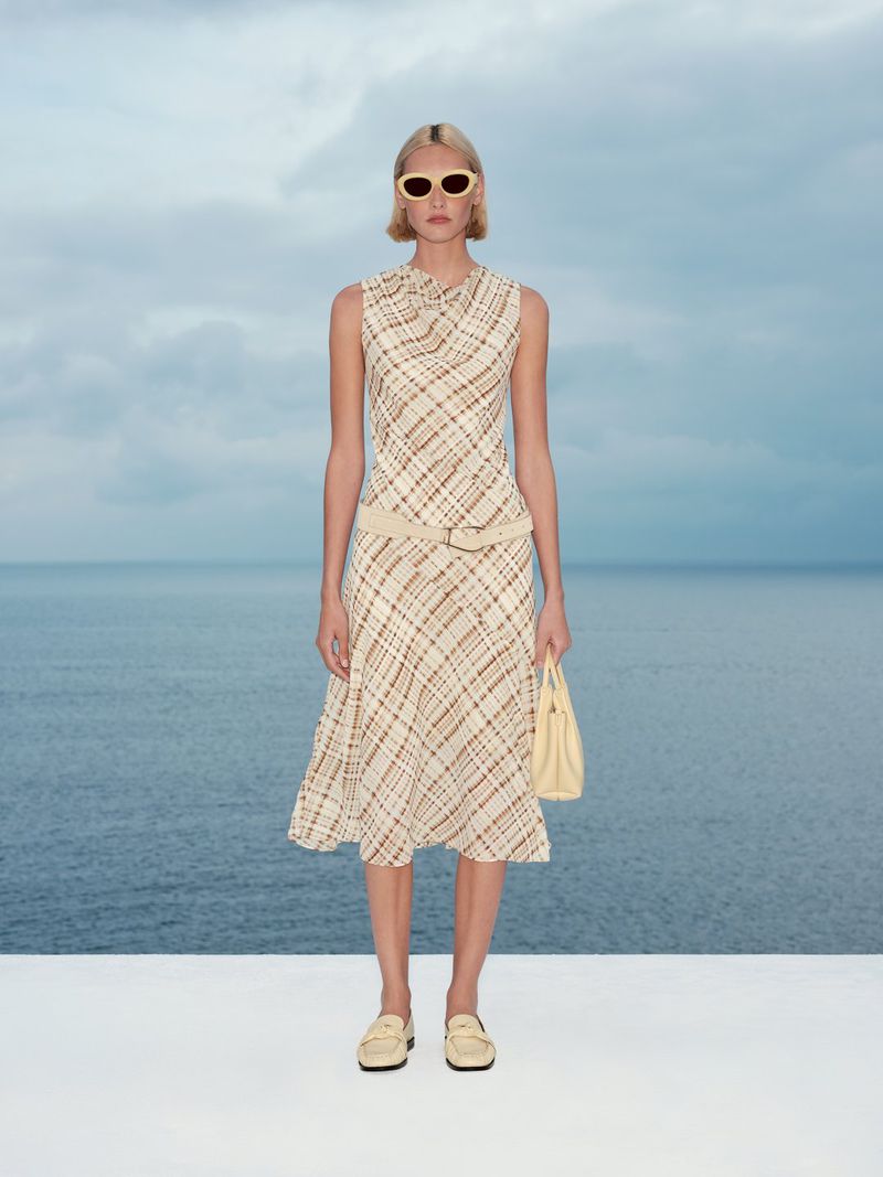 Model wearing patterned dress and sunglasses holding handbag against ocean backdrop