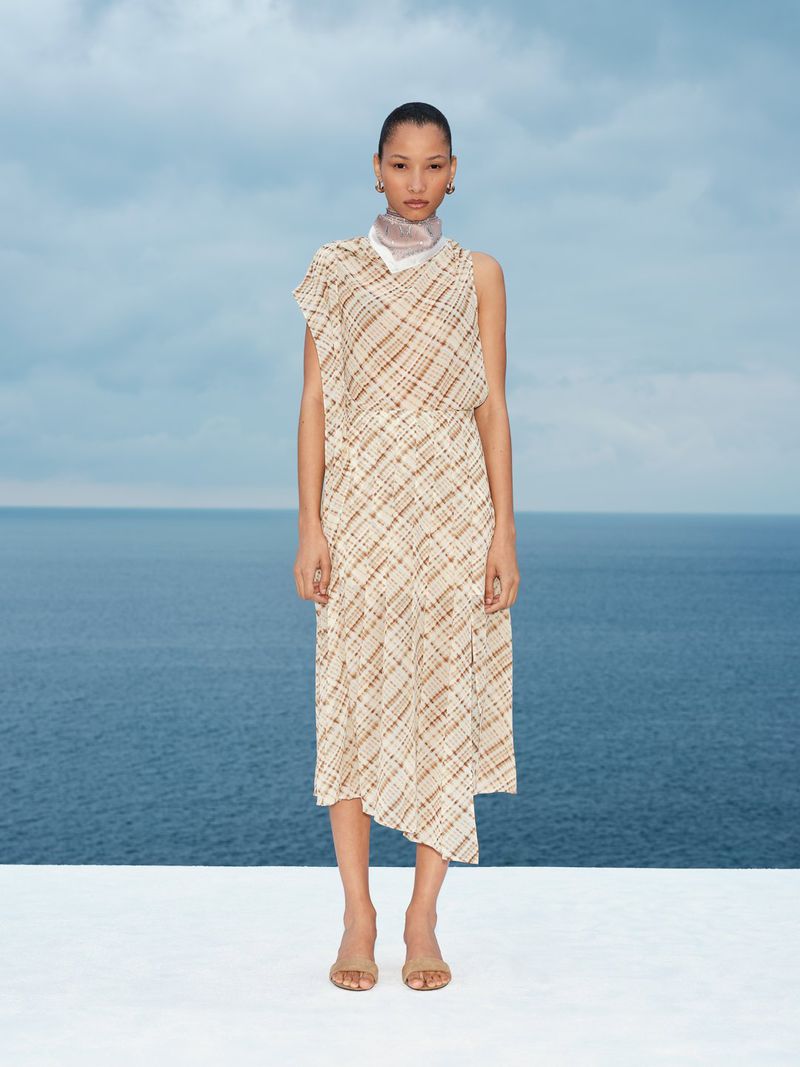 Model standing outdoors wearing a sleeveless patterned midi dress against an ocean and cloudy sky background