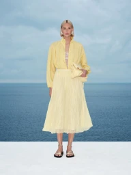Model wearing yellow long-sleeve blouse and pleated skirt holding a clutch bag against a sea view.