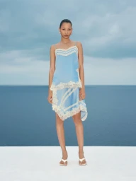 Model wearing a light blue slip dress with white lace trim standing on a platform against an ocean backdrop.