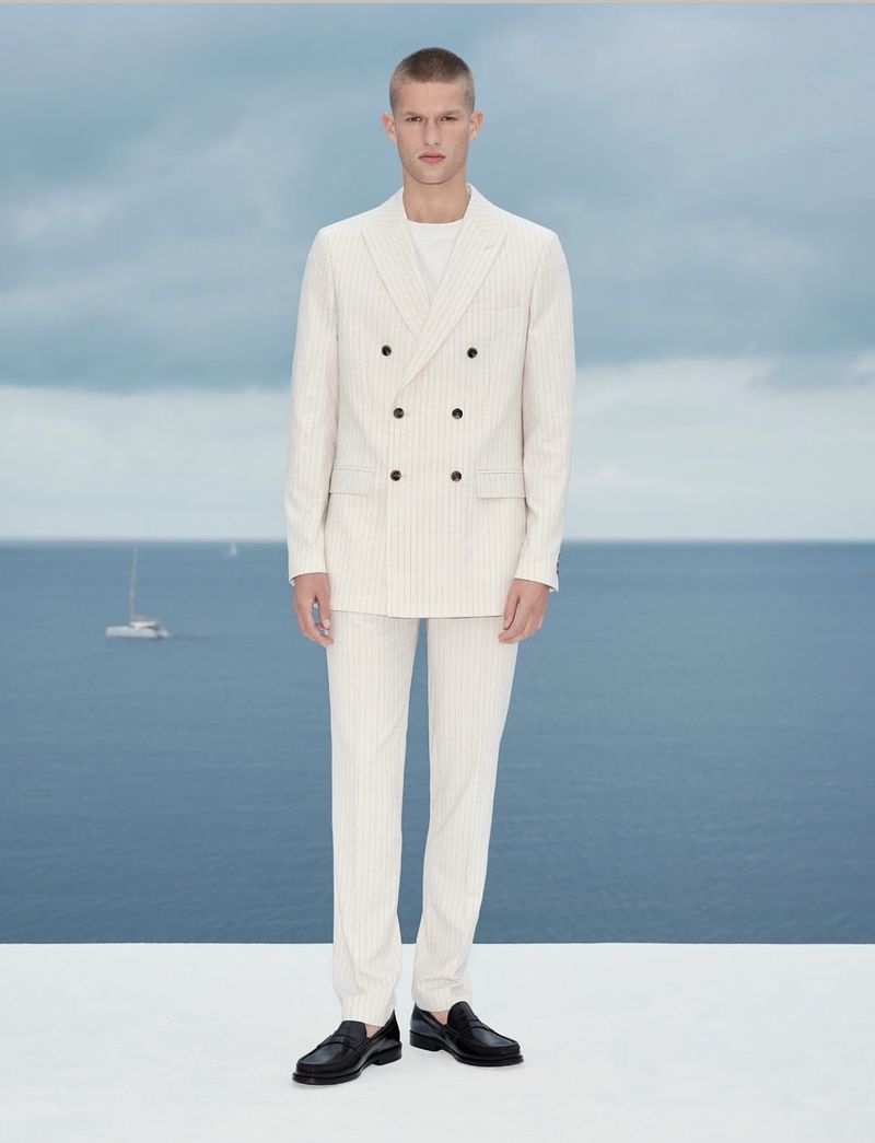 A young fair-skinned man standing in front of the ocean, wearing a double-breasted cream-colored suit and black loafers, posing for a fashion shot.