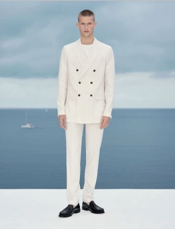 A young fair-skinned man standing in front of the ocean, wearing a double-breasted cream-colored suit and black loafers, posing for a fashion shot.