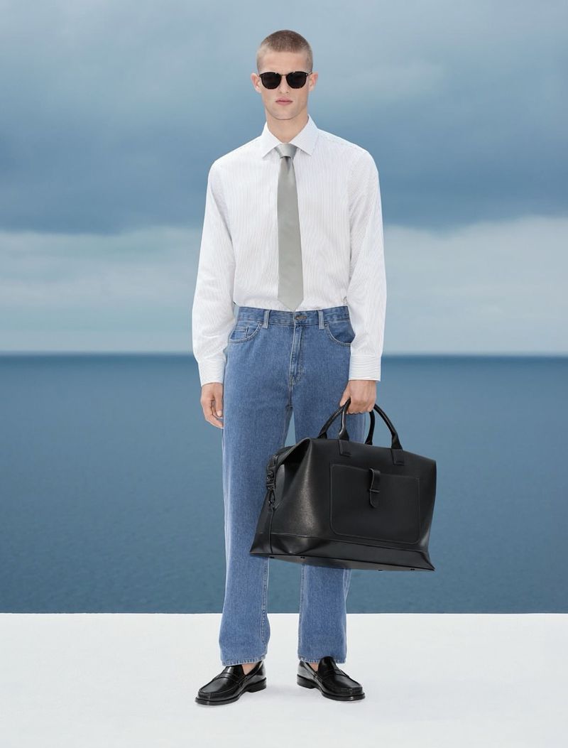 Model wearing a white shirt, tie, and blue jeans holding a black duffel bag against a coastal backdrop.