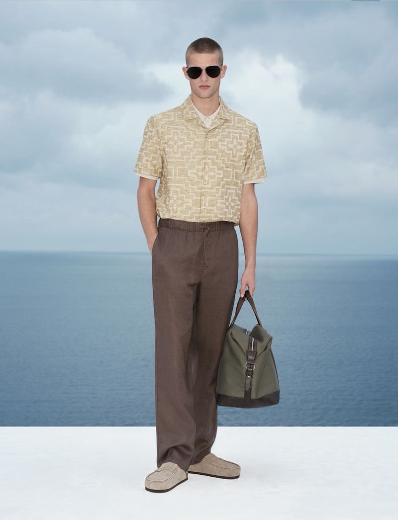 Model wearing patterned shirt, brown trousers, and sunglasses holding a canvas bag against an ocean background