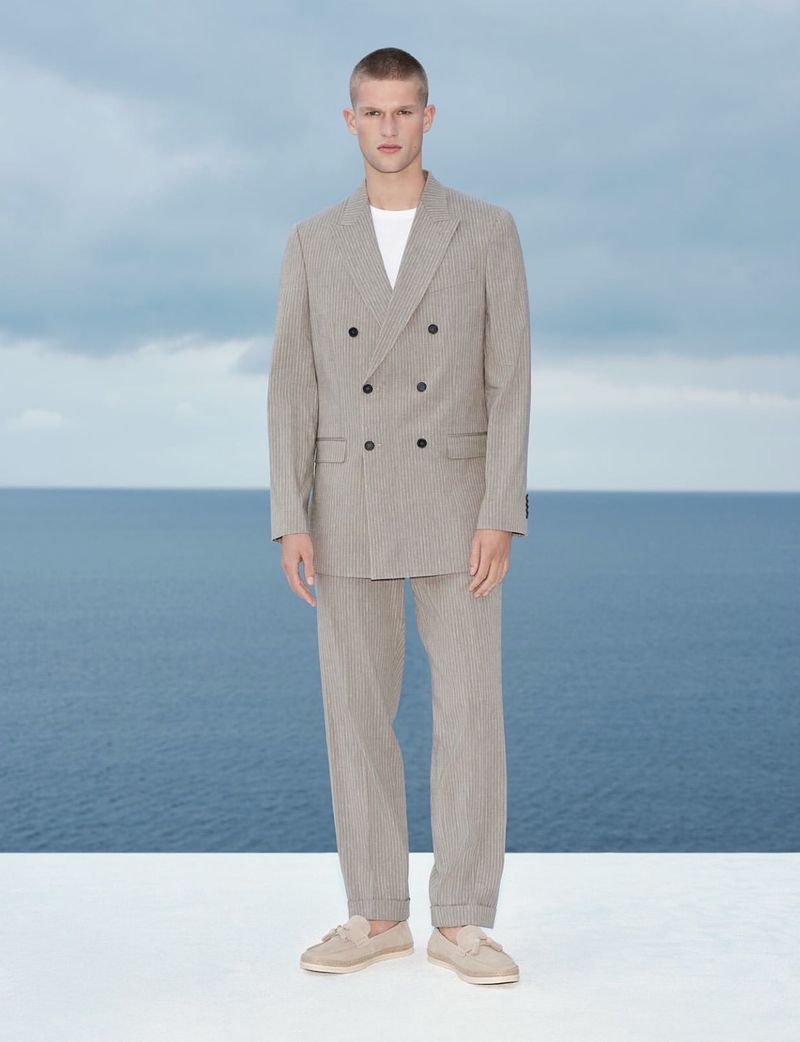 Male model wearing a double-breasted grey suit and light loafers against a coastal sea view backdrop.