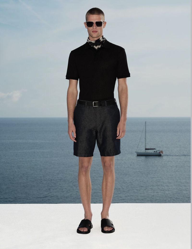 Model in black polo shirt and shorts wearing sunglasses standing against an ocean view backdrop.