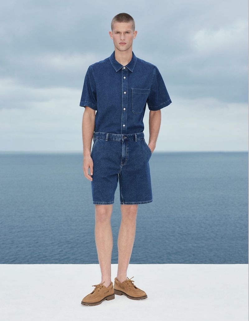 Male model wearing a denim short-sleeve shirt and matching shorts against an ocean and sky backdrop.