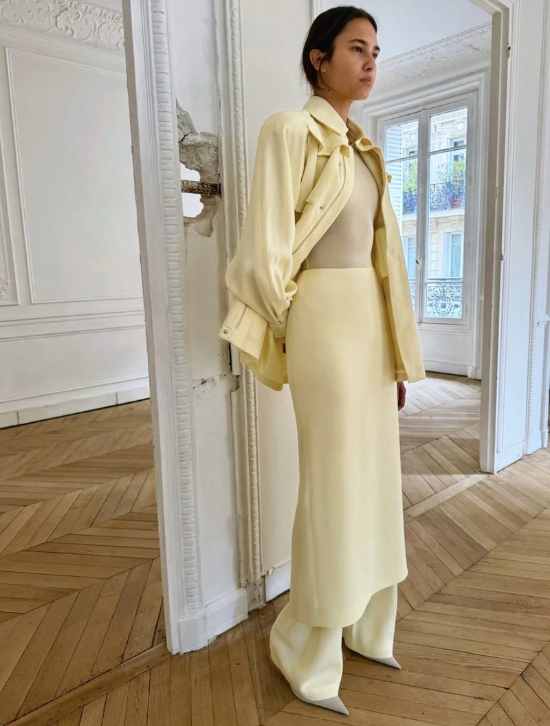 Model in pale yellow suit and neutral top standing in a white Parisian apartment with herringbone flooring.