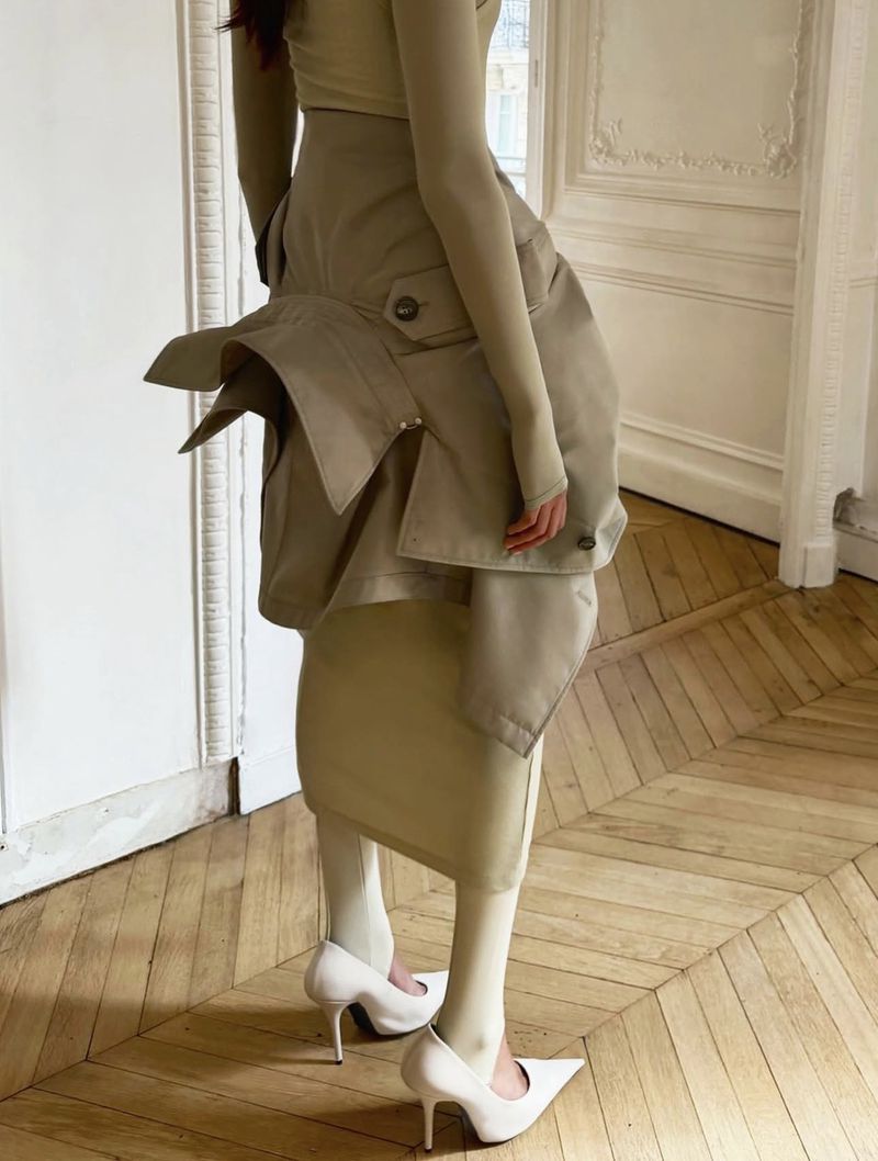 Fashionable woman in beige long-sleeved top, khaki skirt with layered detail, white tights, and white heels posing indoors on a herringbone floor.