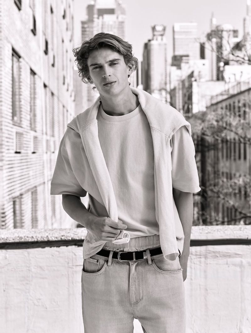 Monochrome fashion portrait of a young man wearing a t-shirt, belted jeans, and another sweater draped over his shoulders.