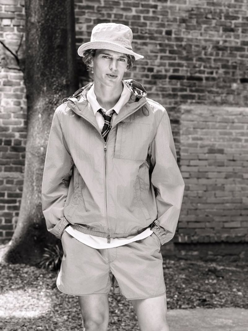Monochrome portrait of a male model wearing a bucket hat, zip jacket, shorts, and tie against a brick wall.