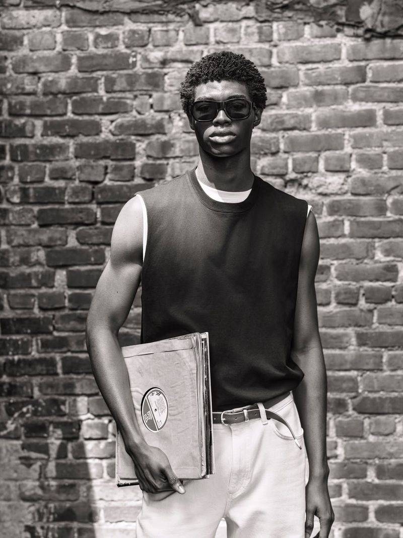 Black man wearing dark sunglasses and a dark sleeveless top holding vinyl records against a brick wall for Zara Origins.