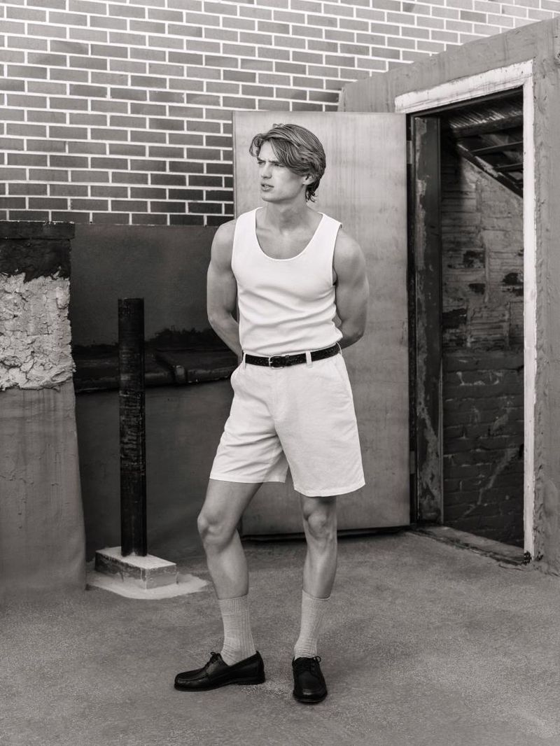 Black and white portrait of a male model wearing a white tank top and shorts against an urban brick building exterior background.