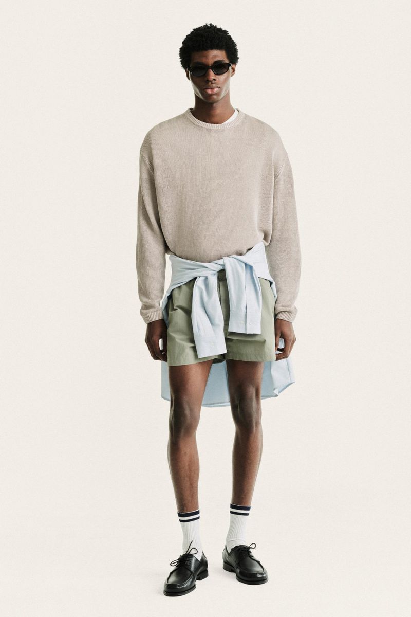 Model standing against a white seamless background wearing a beige crewneck sweater, green shorts, and a light blue shirt tied around the waist.