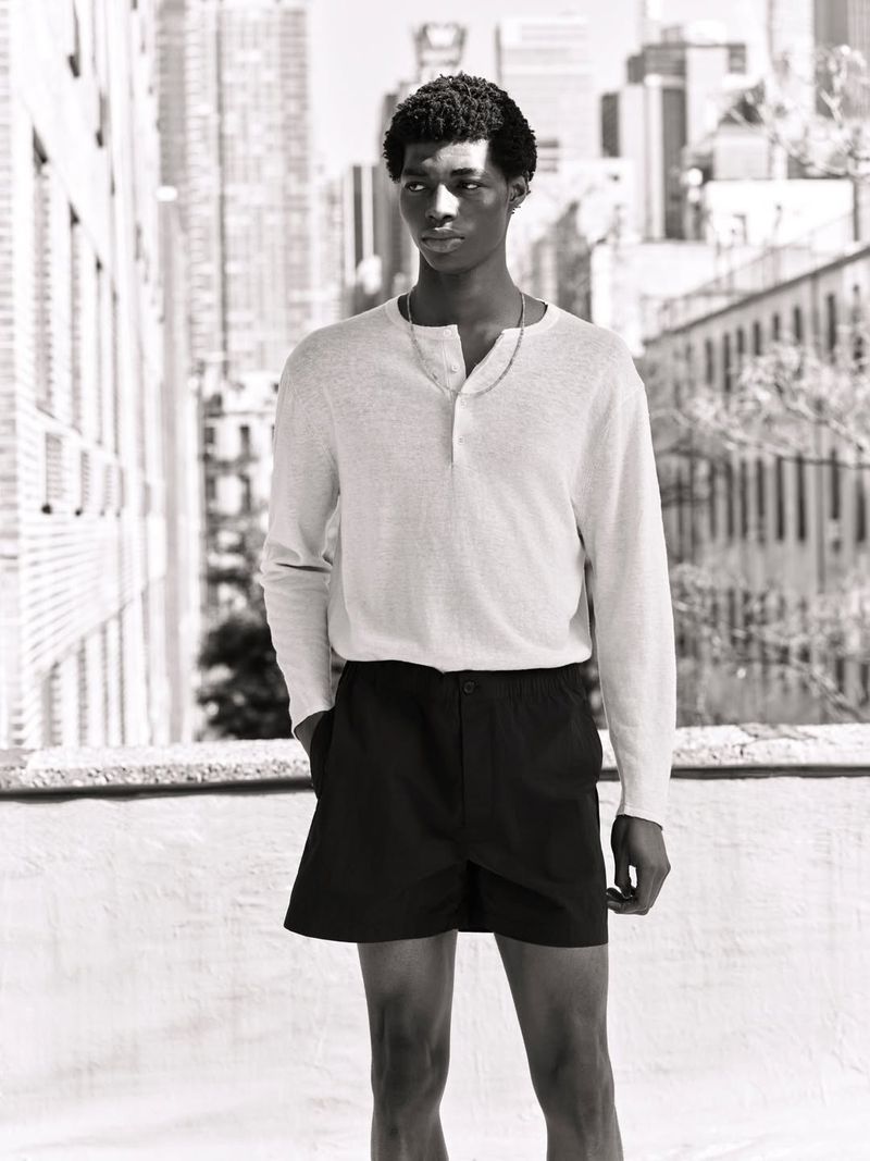 Monochrome portrait of a young man wearing a light henley shirt and dark shorts standing on a city rooftop.