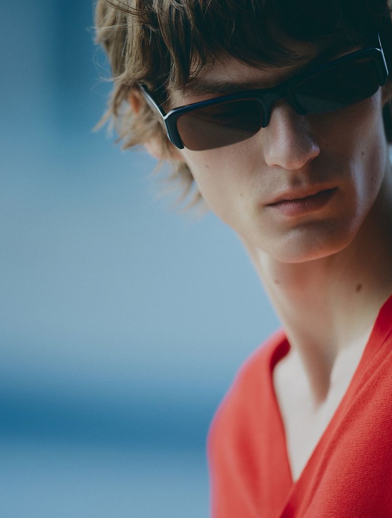 Close-up portrait of a male model wearing black sunglasses and an orange top against a blue background for Zara Man.