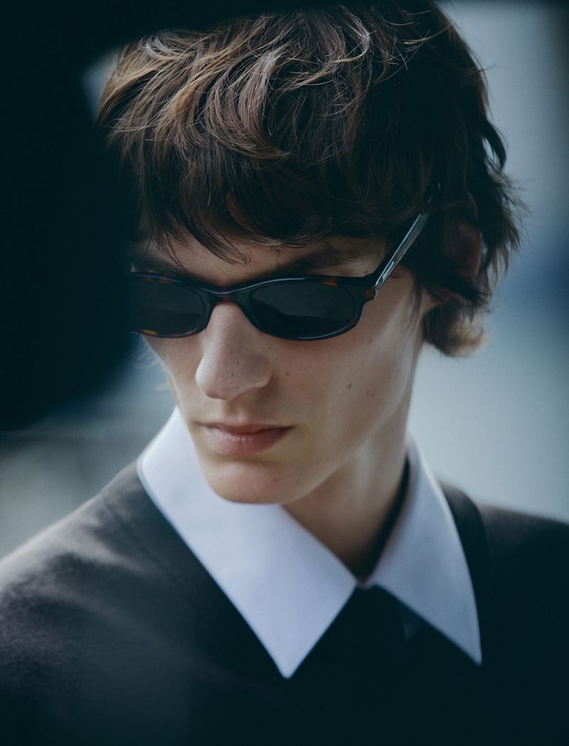 Close-up of a young adult male model with dark hair wearing sunglasses and a dark suit with a white collar, fashionable and stylish.