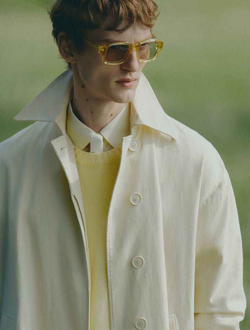 Close-up portrait of a male model wearing tinted yellow sunglasses, a cream shirt, yellow sweater, and light coat.
