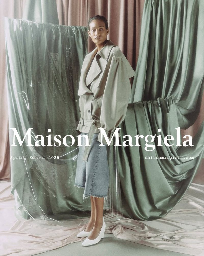 Model standing against draped sage green fabric wearing an oversized beige jacket and denim shorts with white pumps