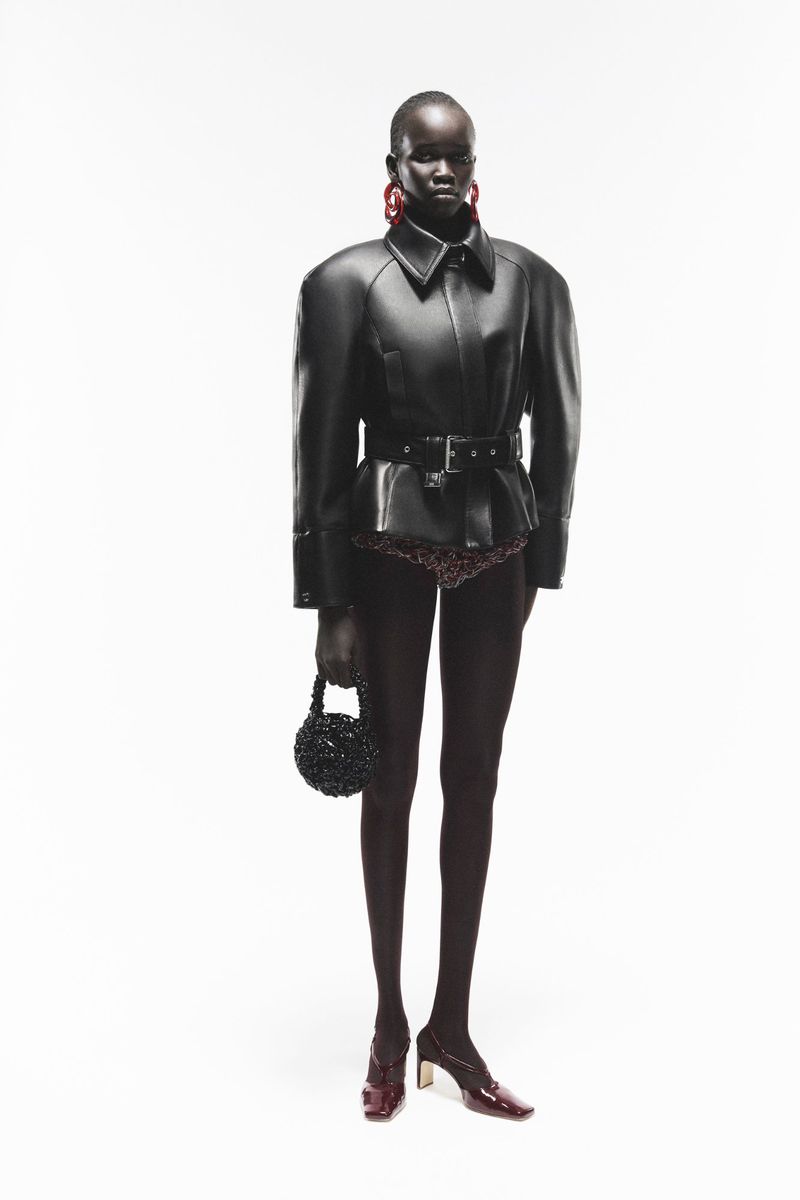 Model wearing a structured black leather belted jacket and holding a small beaded handbag against a white background.