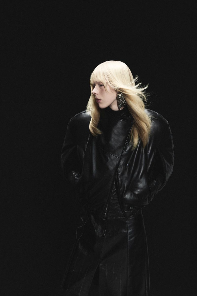 Woman in black leather jacket and pants poses against a dark background with blonde hair and statement earrings, fashion editorial.