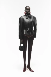 Model wearing a structured black leather belted jacket and holding a small beaded handbag against a white background.