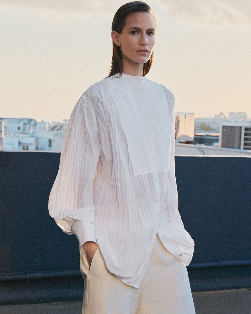 Model in white long-sleeve button-down shirt and cream trousers standing on an outdoor rooftop at sunset