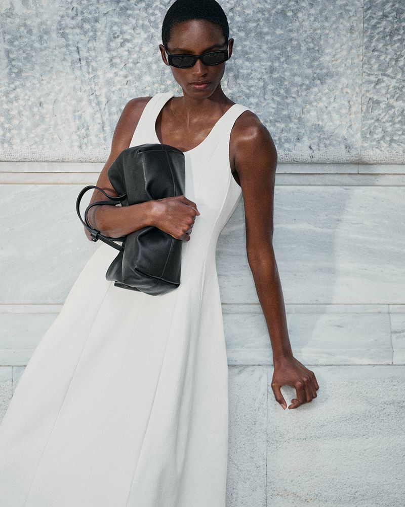 Model in white dress wearing dark sunglasses holding a black leather handbag against a marble wall for COS Summer in the City