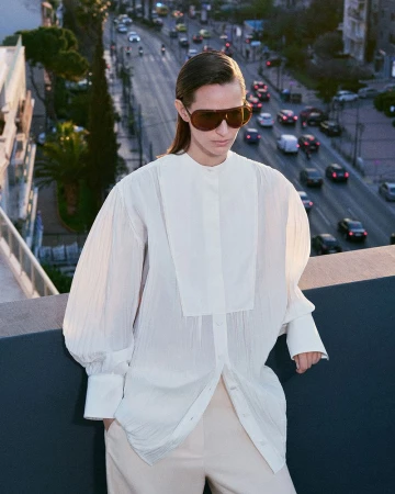 Model wearing dark oversized sunglasses and a white long-sleeve blouse standing on a rooftop overlooking a busy city street.