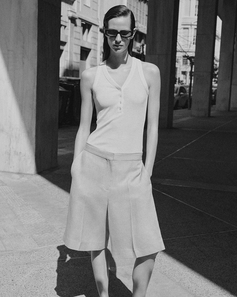 Model in sunglasses wearing a white tank top and tailored shorts outdoors in a city setting