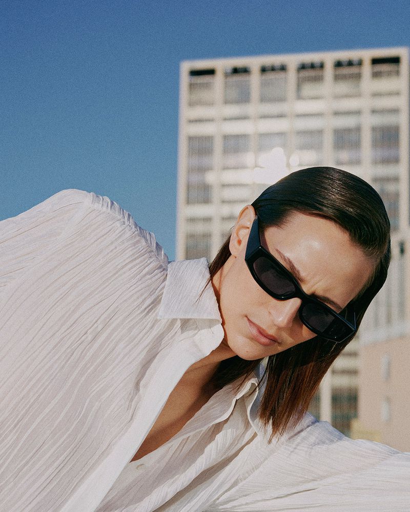Model wearing dark sunglasses and a white textured shirt against a building and blue sky