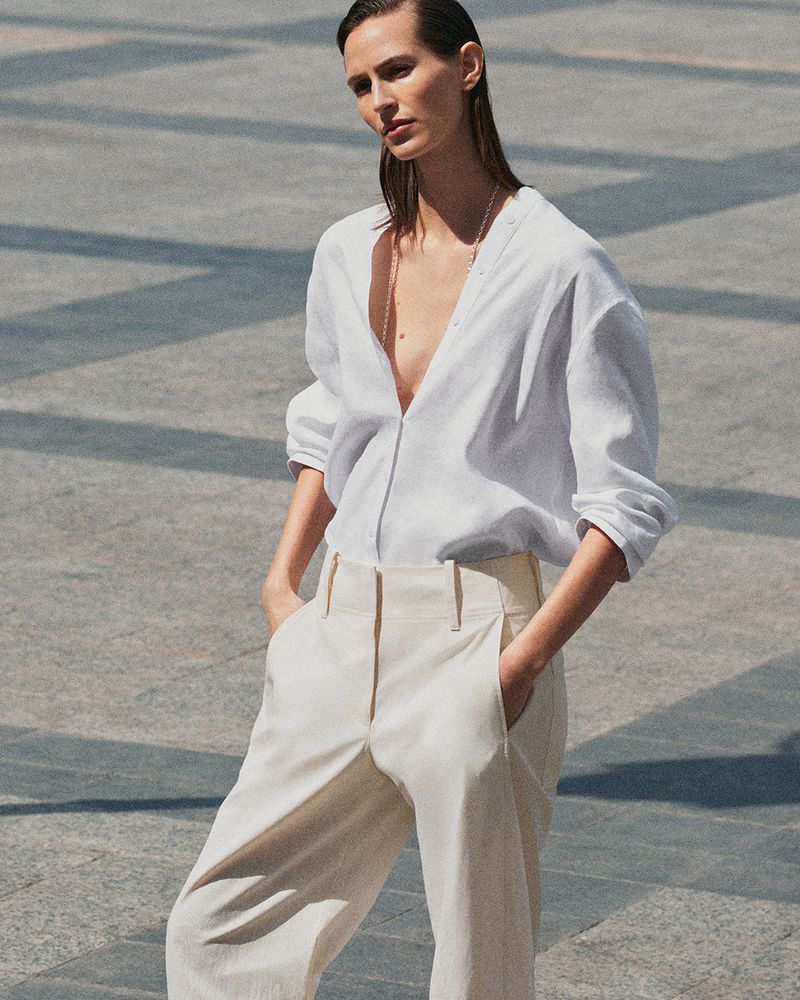 Model wearing a white blouse and light trousers standing outdoors on stone pavement for COS