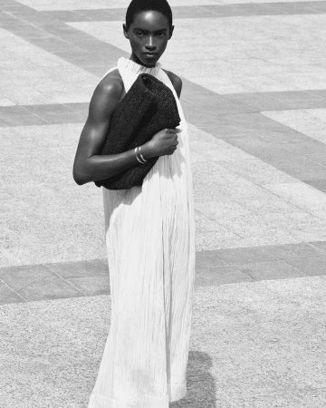 Model in a white pleated maxi dress holding a dark woven clutch on stone paving