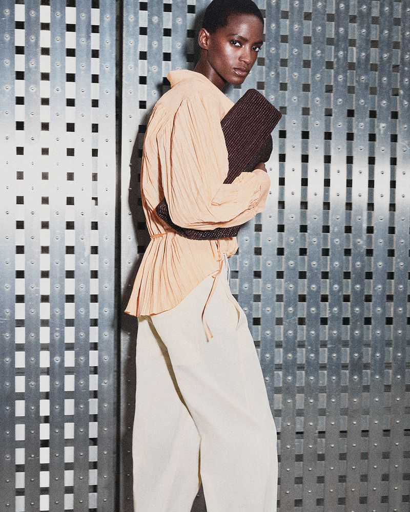 Model wearing a peach blouse and light trousers holding a dark textured clutch against a metal panel background