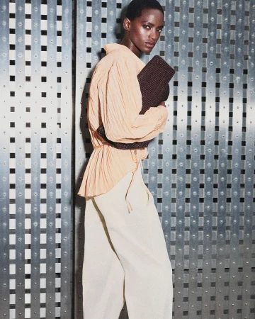 Model wearing a peach blouse and light trousers holding a dark textured clutch against a metal panel background