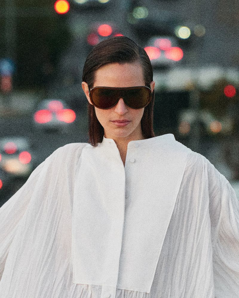 Model wearing dark oversized sunglasses and a white pleated top against an urban street background with bokeh lights