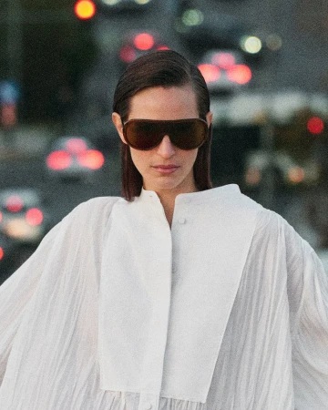 Model wearing dark oversized sunglasses and a white pleated top against an urban street background with bokeh lights
