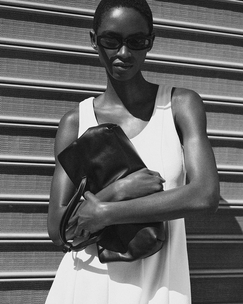 Model in dark sunglasses wearing a white dress and holding a black leather bag against a shuttered storefront
